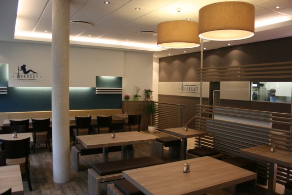 Modernes Restaurant mit hellen Tischen, Holzstühlen und Lampen an der Decke.